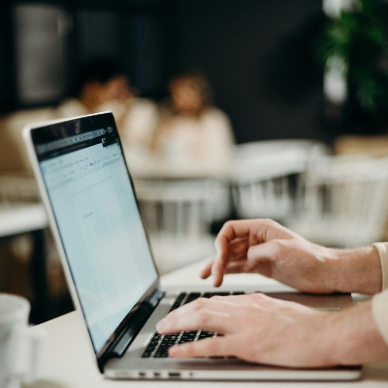 Person typing on a laptop in a cafe with blurred people in the background.