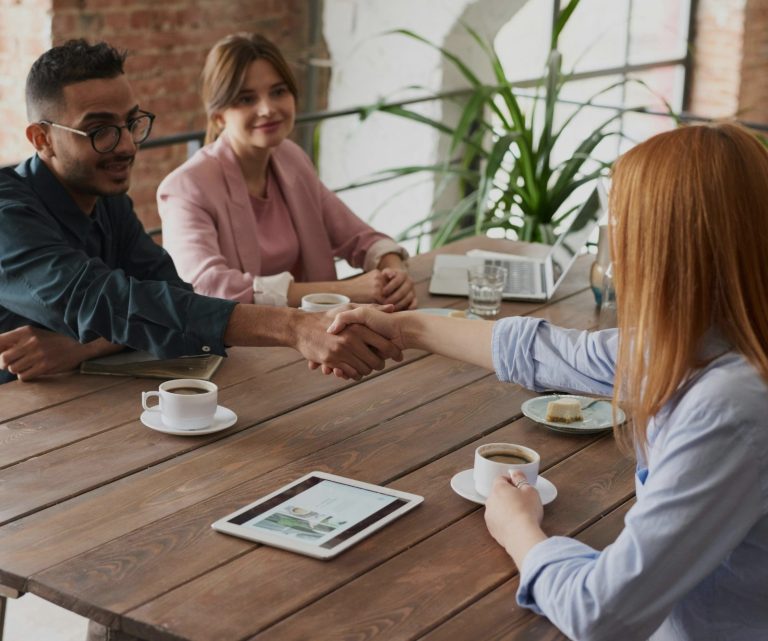 A handshake between two people at a table, with coffee cups and a tablet nearby.